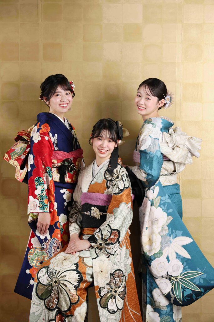 金屏風を背景に、紺・黒・水色の振袖を着た3人の女性が笑顔でポーズをとる成人式記念写真